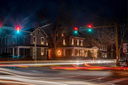A nighttime street scene with traffic lights, illuminated houses, and light trails from passing cars.