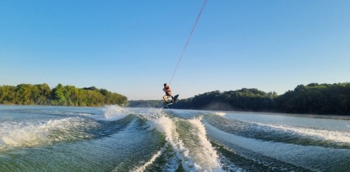 A person wakesurfing on a calm lake, jumping off a wave with a clear blue sky and trees in the background.