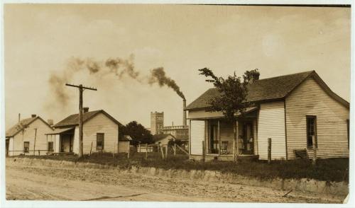Historic sepia-toned image of houses along a dirt road, with smoke rising from a factory in the background.