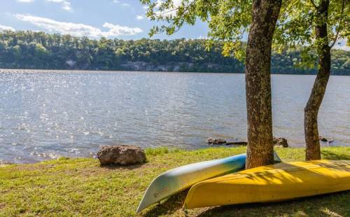 Two canoes rest on the grass by a calm lake, surrounded by trees and a scenic view of the shoreline.