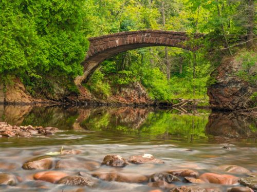 A stone bridge arches over a calm river, surrounded by lush green trees and rocky riverbed reflections.
