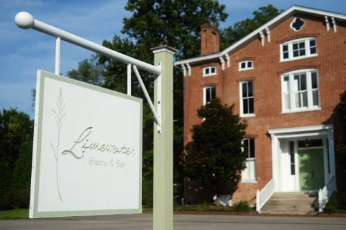 Sign for Limeswater Bistro & Bar in front of a historic brick building surrounded by trees. Clear blue sky above.
