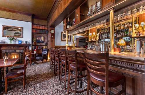 A cozy bar with wooden stools, a polished counter, and shelves lined with bottles in a warmly lit setting.