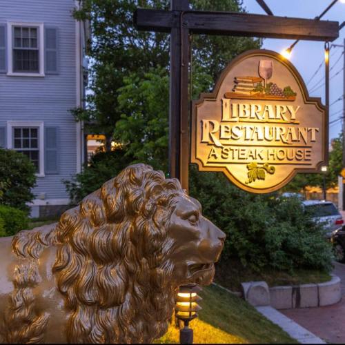 A golden lion statue in front of the Library Restaurant sign, a steakhouse, with greenery and buildings in the background.