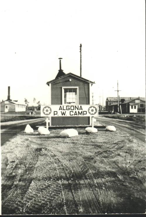 Sign for Algona P.W. Camp, with a building and gravel road in a historical black-and-white photograph.