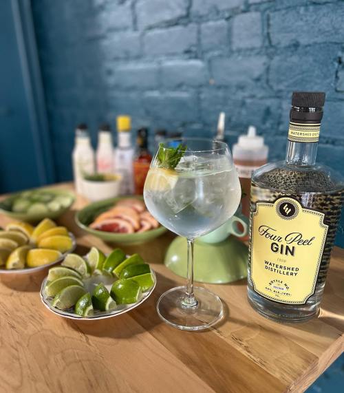 A glass of gin with mint sits on a wooden table, surrounded by fresh limes and other cocktail ingredients.