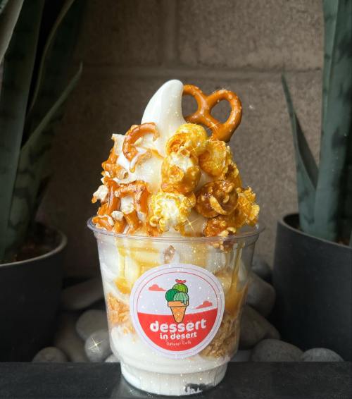 A cup of soft serve ice cream topped with caramel popcorn, pretzels, and a sprinkle of crushed cookies.