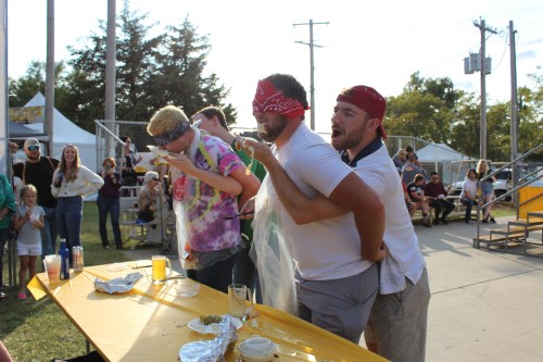 Three blindfolded participants compete in a messy eating contest at an outdoor event, with spectators watching nearby.
