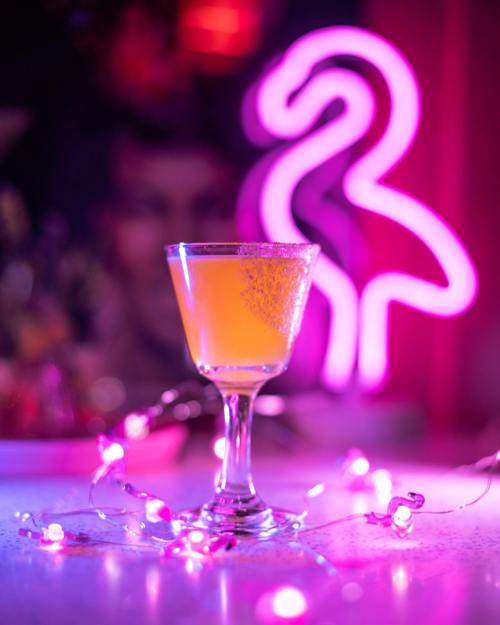 A vibrant cocktail in a glass, surrounded by pink fairy lights and a neon flamingo sign in the background.