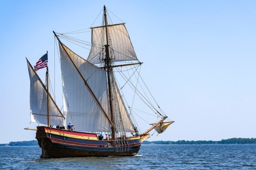 A historic sailing ship with billowing sails and an American flag, gliding across calm waters under a clear blue sky.