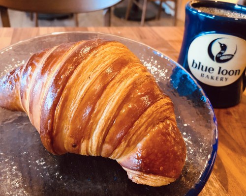 A golden-brown croissant on a glass plate next to a blue mug labeled "blue loon Bakery."