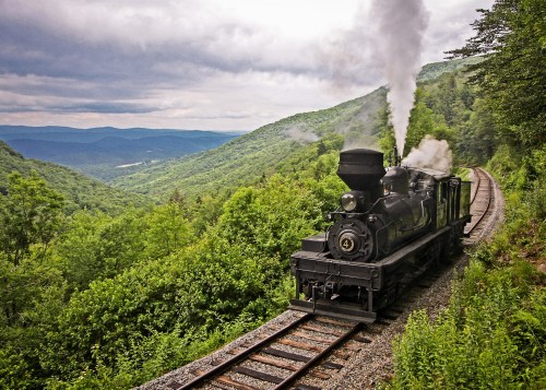 A vintage steam train chugs along a winding track through lush green mountains under a cloudy sky.