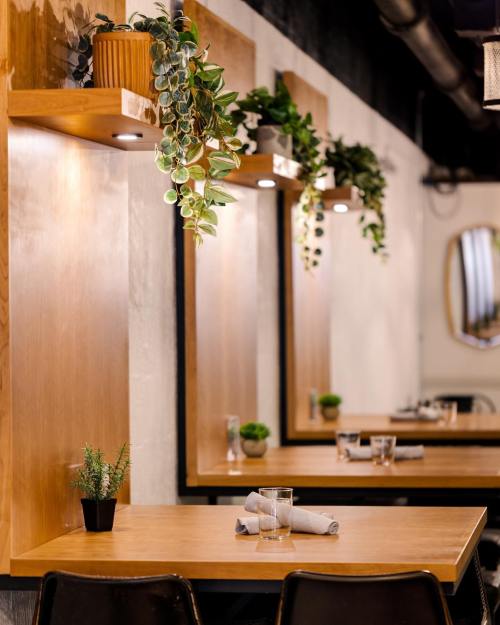 A cozy restaurant interior with wooden tables, plants on shelves, and soft lighting.