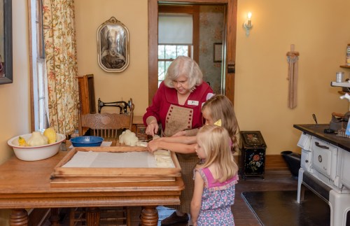 An elderly woman teaches two young girls how to roll dough in a cozy kitchen.