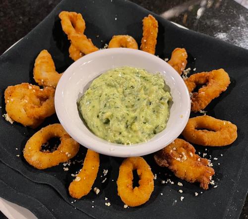 A plate of fried calamari rings arranged around a bowl of green dipping sauce on a black napkin.