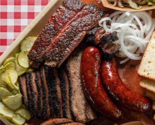 A platter of barbecue featuring ribs, brisket, sausage, pickles, onions, and slices of bread on a checkered tablecloth.