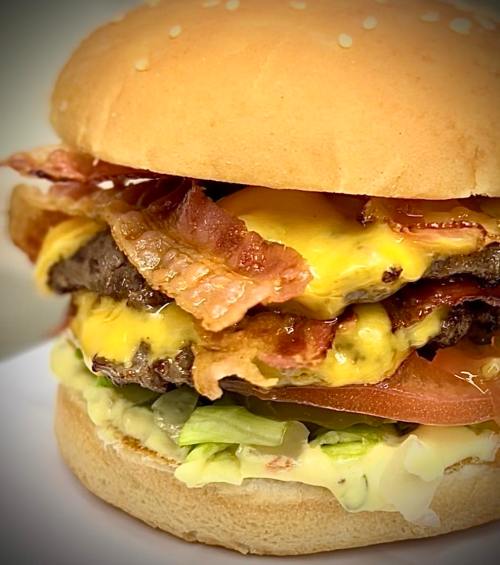 A close-up of a juicy burger with two beef patties, melted cheese, bacon, lettuce, tomato, and mayonnaise on a sesame seed bun.