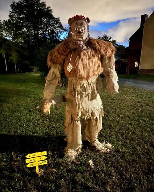 A large, scarecrow-like figure resembling a bear, made of straw and fabric, stands in a dimly lit outdoor setting.