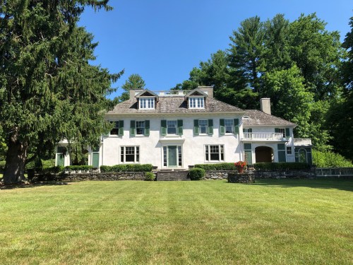A large, white two-story house with green shutters, surrounded by trees and a grassy lawn under a clear blue sky.
