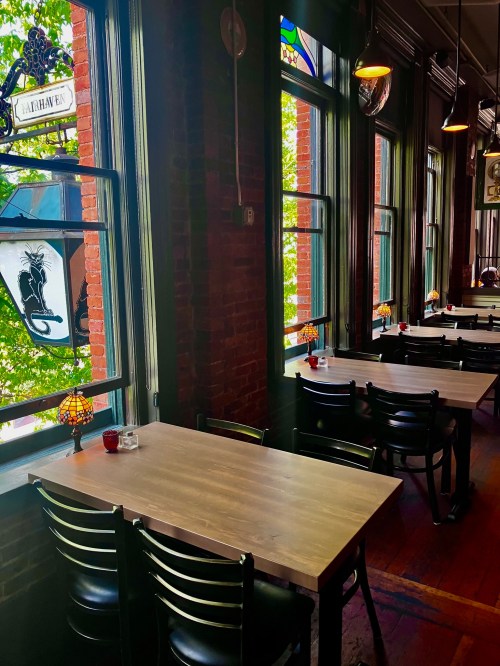 Bright restaurant interior with wooden tables, black chairs, and large windows showcasing greenery outside.