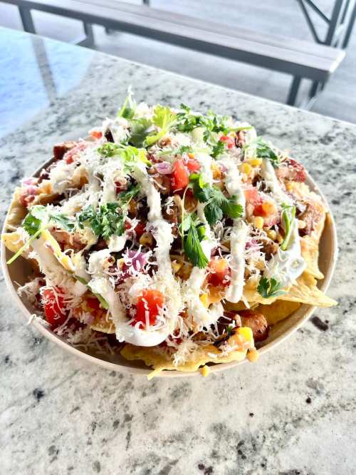 A bowl of nachos topped with cheese, tomatoes, onions, cilantro, and drizzled with sauce on a marble surface.