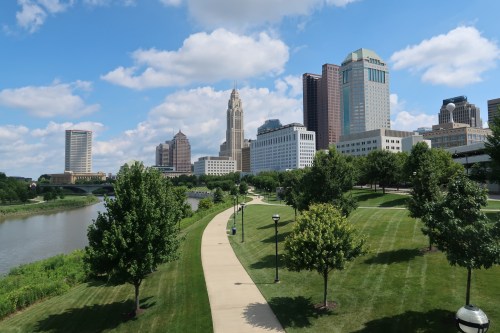 A scenic view of a city skyline with green parks and a river under a blue sky with fluffy clouds.