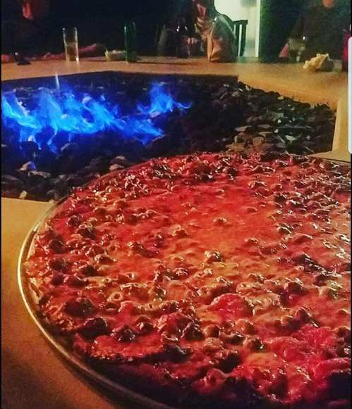 A close-up of a pizza with toppings, placed near a glowing blue fire pit in a dimly lit setting.