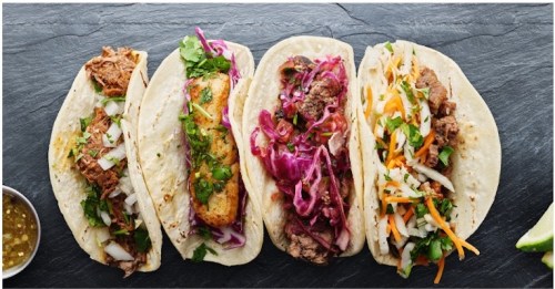 Four delicious tacos on a slate platter, filled with various meats, toppings, and garnished with fresh herbs.
