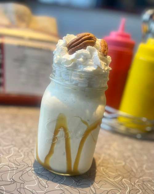 A mason jar filled with a creamy milkshake topped with whipped cream and pecans, drizzled with caramel sauce.