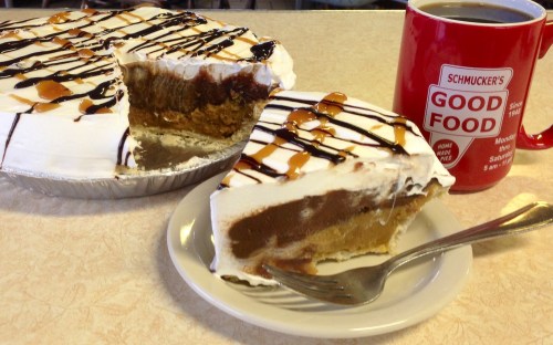 A slice of pie with whipped cream and chocolate drizzle on a plate, next to a mug of coffee.