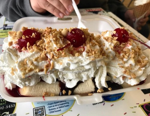 A dessert with whipped cream, chopped nuts, cherries, and bananas, served in a takeout container.