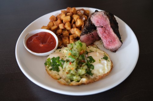 A plate with sliced steak, fried potatoes, a poached egg topped with salsa, and a small bowl of ketchup.