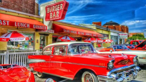 A vintage red car parked outside a retro diner with umbrellas and a bright blue sky in the background.