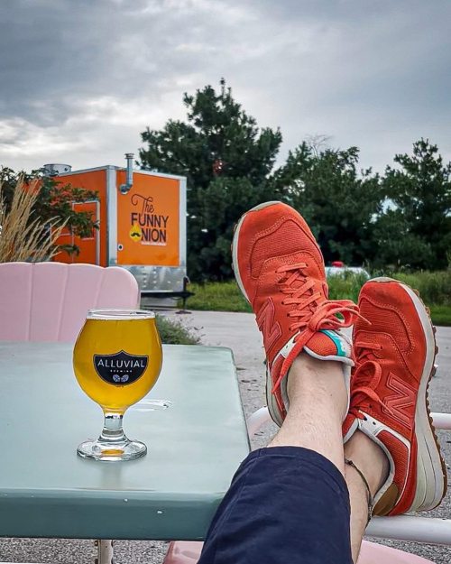 A person relaxes outdoors with orange sneakers on a table next to a glass of beer, with a food truck in the background.