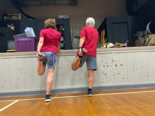 Two people in red shirts stretch their legs while facing a wall in a gym setting.