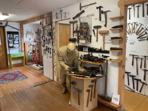 A sculpted figure examines tools on a table in a workshop filled with various hand tools on the walls.