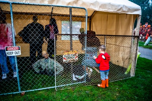 A child in a red hoodie and orange boots stands by a fenced area with spooky decorations and mannequins.