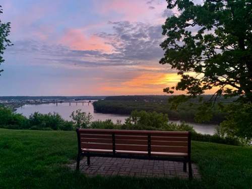 A wooden bench overlooks a river at sunset, with colorful clouds and greenery surrounding the scene.