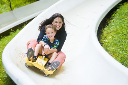 A woman and a young boy joyfully ride down a winding slide surrounded by greenery.