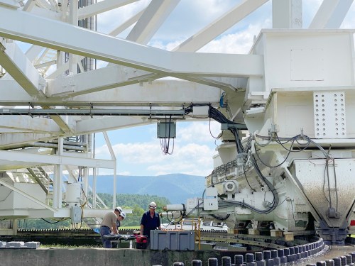 Two workers inspect machinery on a large structure, with mountains and a blue sky in the background.