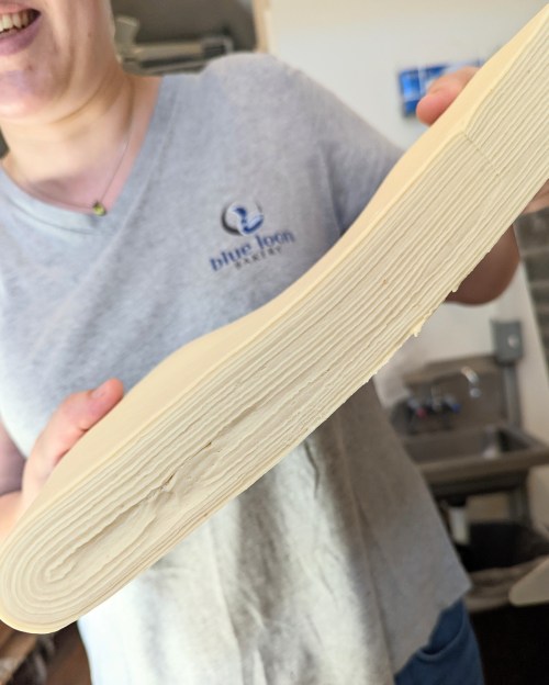A person holds a thick, layered piece of dough, smiling in a kitchen setting.