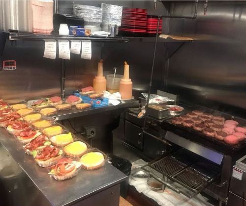 A busy kitchen with burgers grilling and trays of prepared food, including burgers and toppings, on the counter.