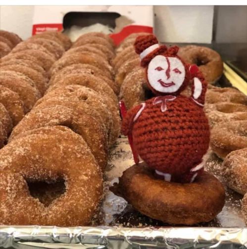 A crocheted figure stands on a donut, surrounded by a tray of cinnamon sugar donuts.