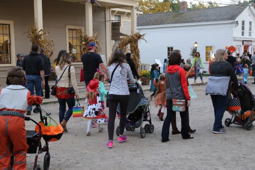 A crowd of families and children in costumes walking down a dirt path, with strollers and festive decorations around.