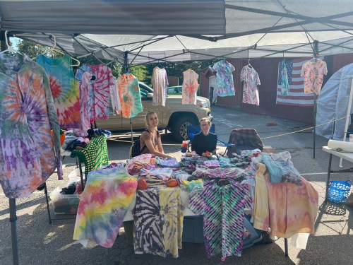 Two people sit at a tie-dye clothing booth, surrounded by colorful shirts and a backdrop of an American flag.