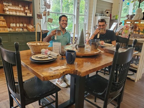 Two men sit at a rustic wooden table in a cozy shop, each holding a mug and smiling. Decor and natural light surround them.
