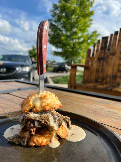 A biscuit sandwich with beef and creamy sauce, topped with a knife, on a wooden table with a blurred background.