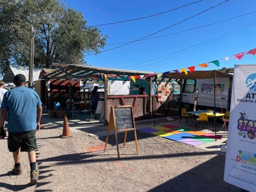 A colorful outdoor market with tents, tables, and a sign, featuring people and decorative banners.