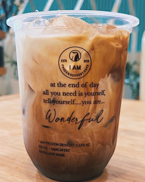 A clear cup of iced coffee with a motivational quote printed on it, placed on a wooden table.