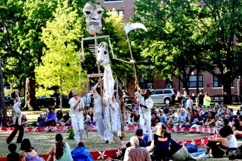 A group of performers manipulate a large puppet in a park, surrounded by an audience seated on the grass.